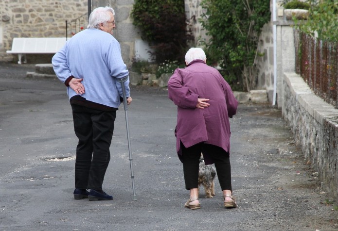 Two older adults, one using a cane, walk with a small dog, illustrating mobility challenges and the need for dog walking assistance in assisted living.